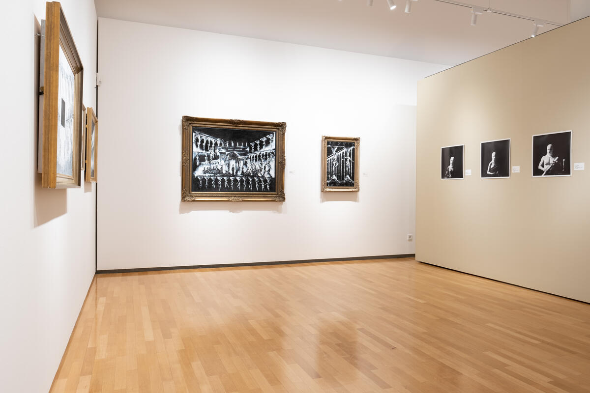 Bild vergrößern: Ausstellungsraum mit in Goldrahmen präsentierter Malerei antiker Stätten und ungerahmten Schwarz-Weiß-Fotografien an den Wänden.