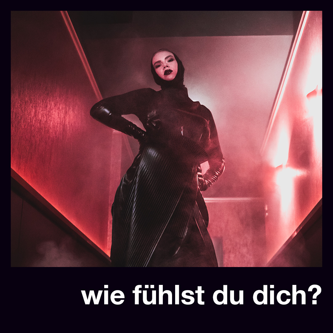 Bild vergrößern: Ein Model posiert in einem schwarzen Kleid in einem rot beleuchteten Raum. Darunter steht die Frage: Wie fühlst du dich?