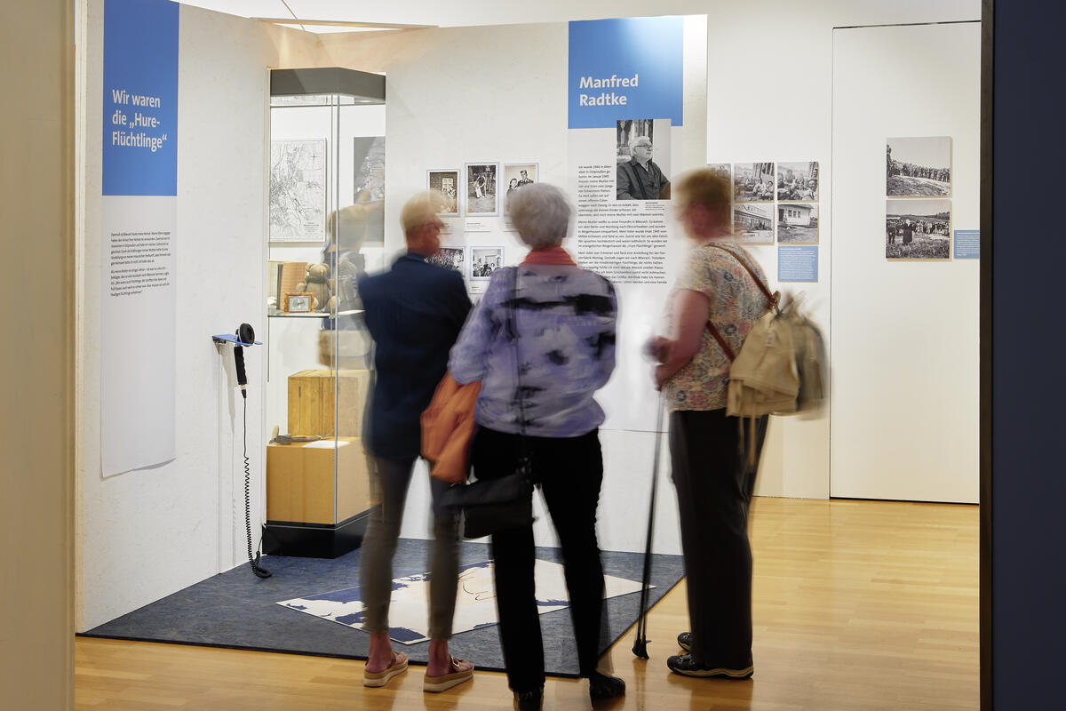 Bild vergrößern: Ausstellungsraum mit Besuchern, die die Geschichte des Zeitzeugens Manfred Radtke anschauen.