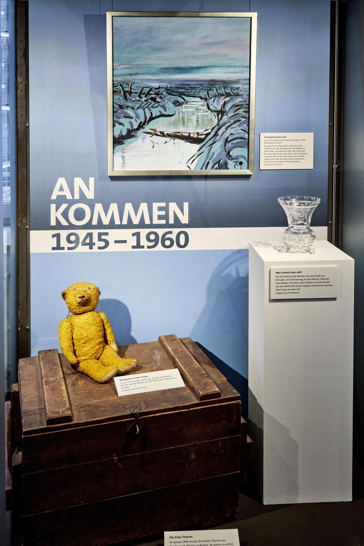 Bild vergrößern: Ausstellungsraum mit einem Teddy, einer Holzkiste und einer Kristallvase, die Menschen auf der Flucht mitgenommen haben.