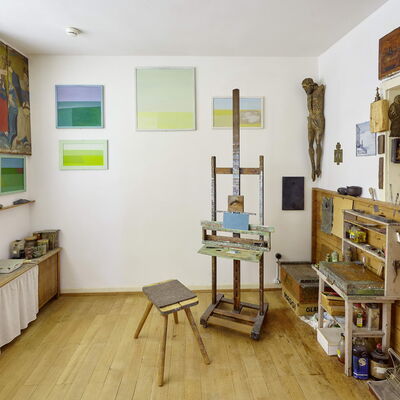 Bild vergrößern: Eine Staffelei steht vor eiiner mit Kunstwerken und Objekten behängten Wand.